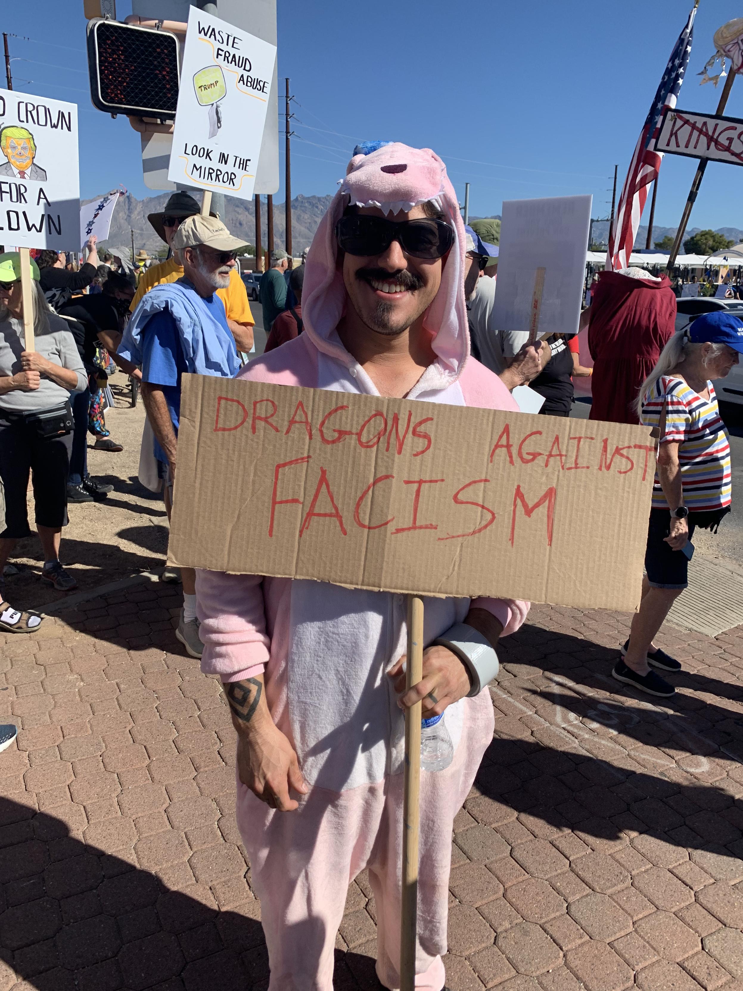 Man dressed in a pink dragon onesie holding a sign "dragons against fascism"