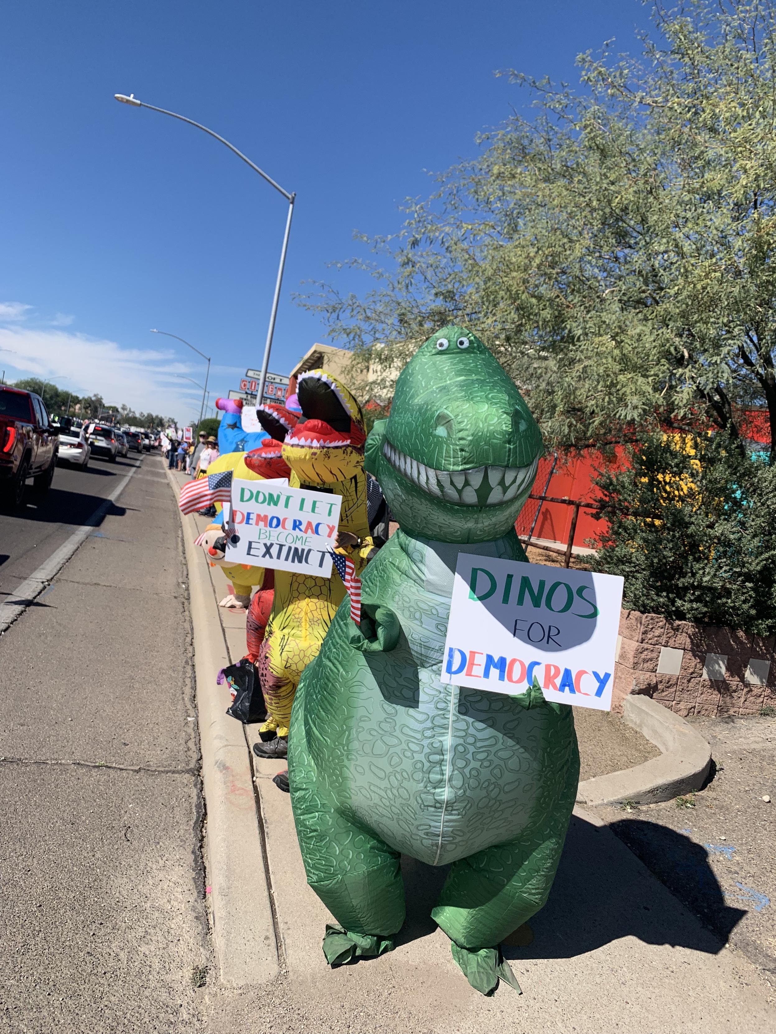 People in inflatable dinosaur costumes along a sidewalk, with signs "Dinos for democracy" and "Don't let democracy become extinct"