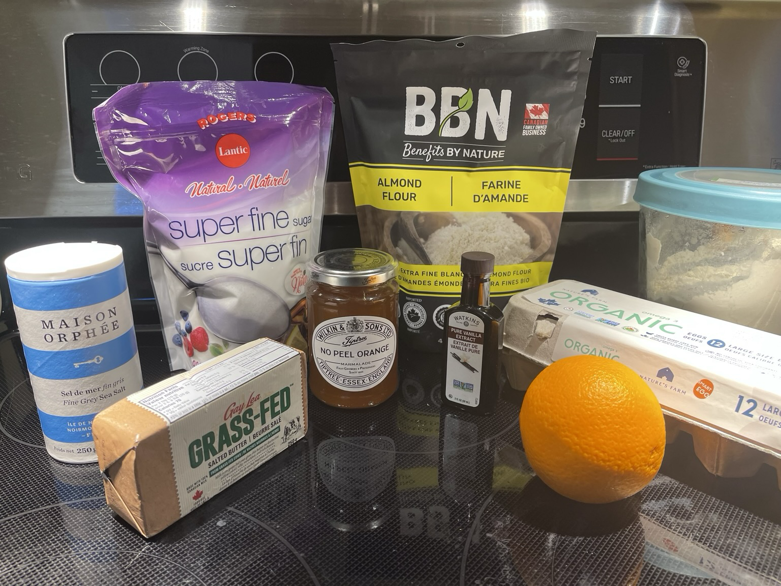 Ingredients for the EBTSC weekend attempt at the Guardian Breton butter cake recipe on a flat black glass stovetop.  From left to right: Maison Orphée grey sea salt; Gay Lea grass fed butter with sea salt; Lantic superfine sugar; Wilkin & Sons no-peel marmalade; Watkins Madagascar vanilla extract; BBN almond flour; a large orange; a carton of organic eggs; and a tall glass container with a sky blue lid containing flour.