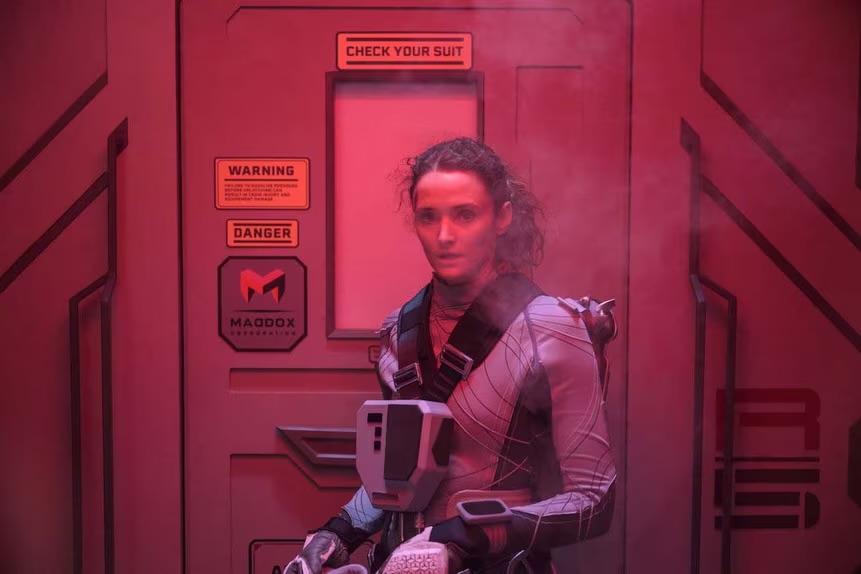 The Ark S1E10: Lt. Garnett stands in an airlock with her back to the hatch.  She is tallish and light completed.  She is wearing a gray environmental suit but no helmet.  Her long curly hair is pulled back in a ponytail.  There is an instrumentation box strapped to her chest, with large black harness straps running back over her shoulders.  The lock is illuminated with red dim light.  Her expression is pensive as she looks forward to something or someone offscreen.